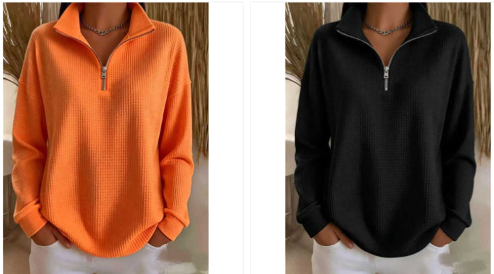 Two women modeling the same quarter-zip pullover sweater in different colors: one bright orange on the left and one black on the right. Both are shown from neck to hips with hands in white pants, against a neutral indoor backdrop with dried grass decor.