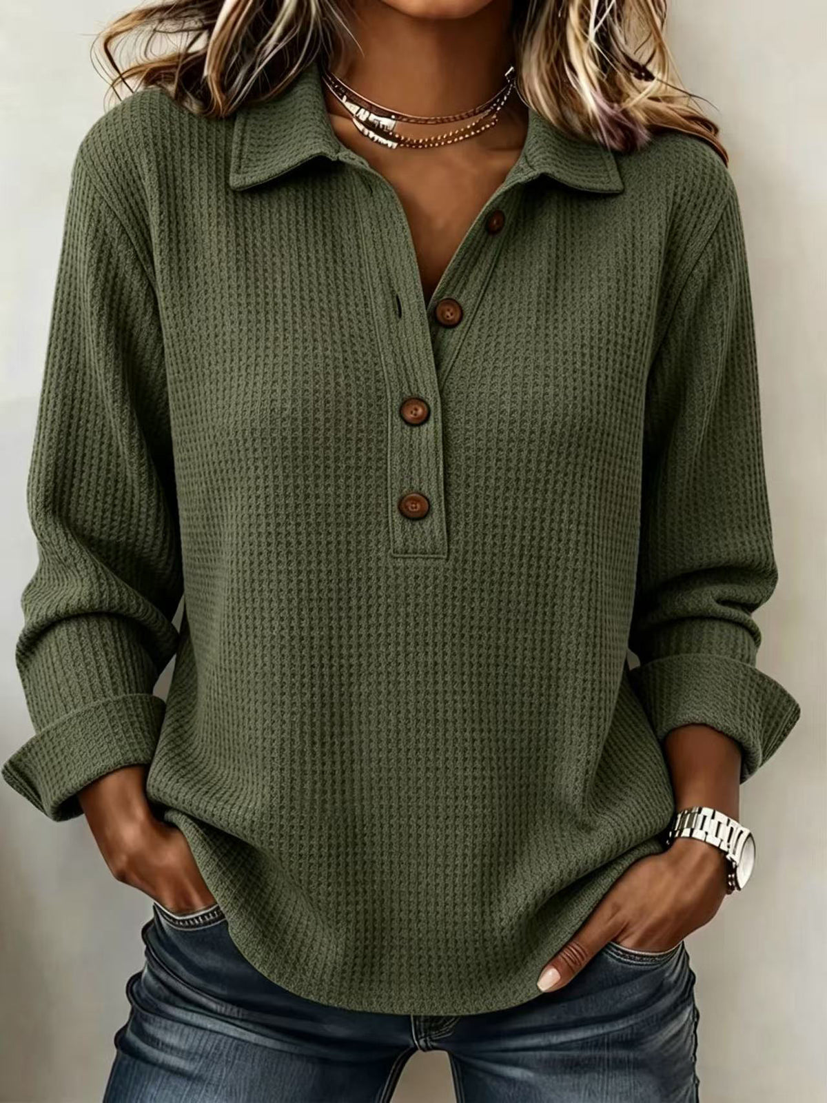 A woman wearing an olive green waffle-knit long-sleeve polo shirt with brown buttons, paired with blue jeans and layered necklaces.