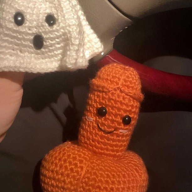 A close-up of a hand holding a small orange crocheted plush figure with a smiling face, next to a white crocheted ghost-like item.