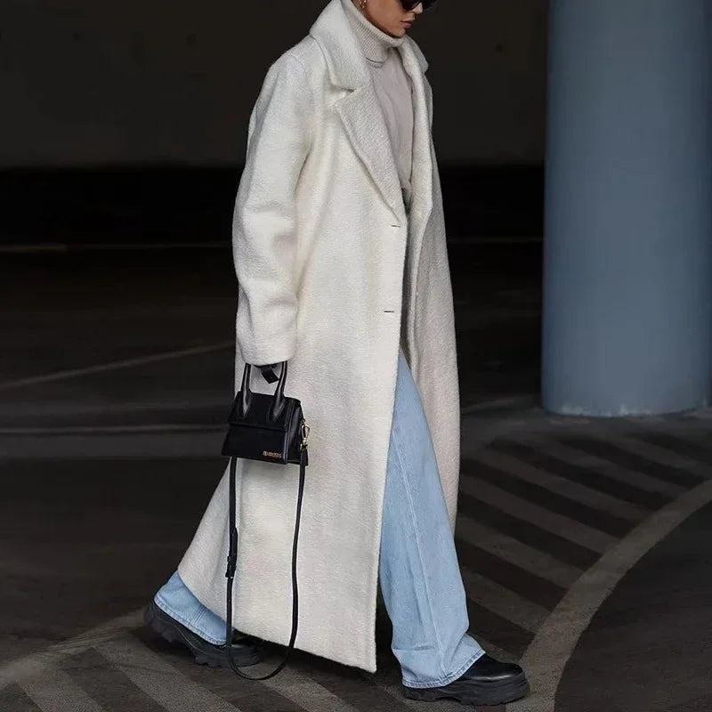 Woman in a long white wool coat, turtleneck sweater, light blue jeans, and chunky black boots holding a black handbag, showcasing chic winter street style.