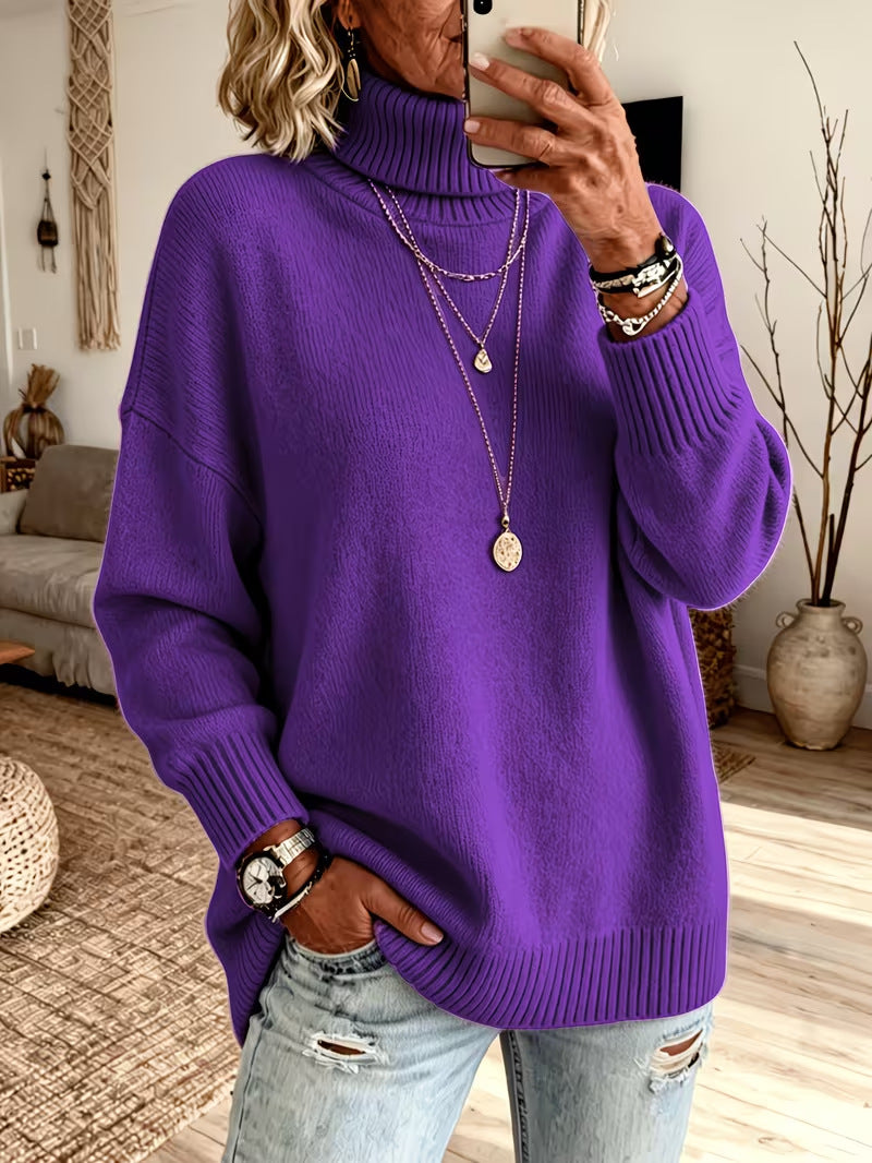 A woman takes a mirror selfie wearing an oversized purple ribbed turtleneck sweater, light-wash distressed jeans, layered gold necklaces, and bracelets in a cozy living room setting.