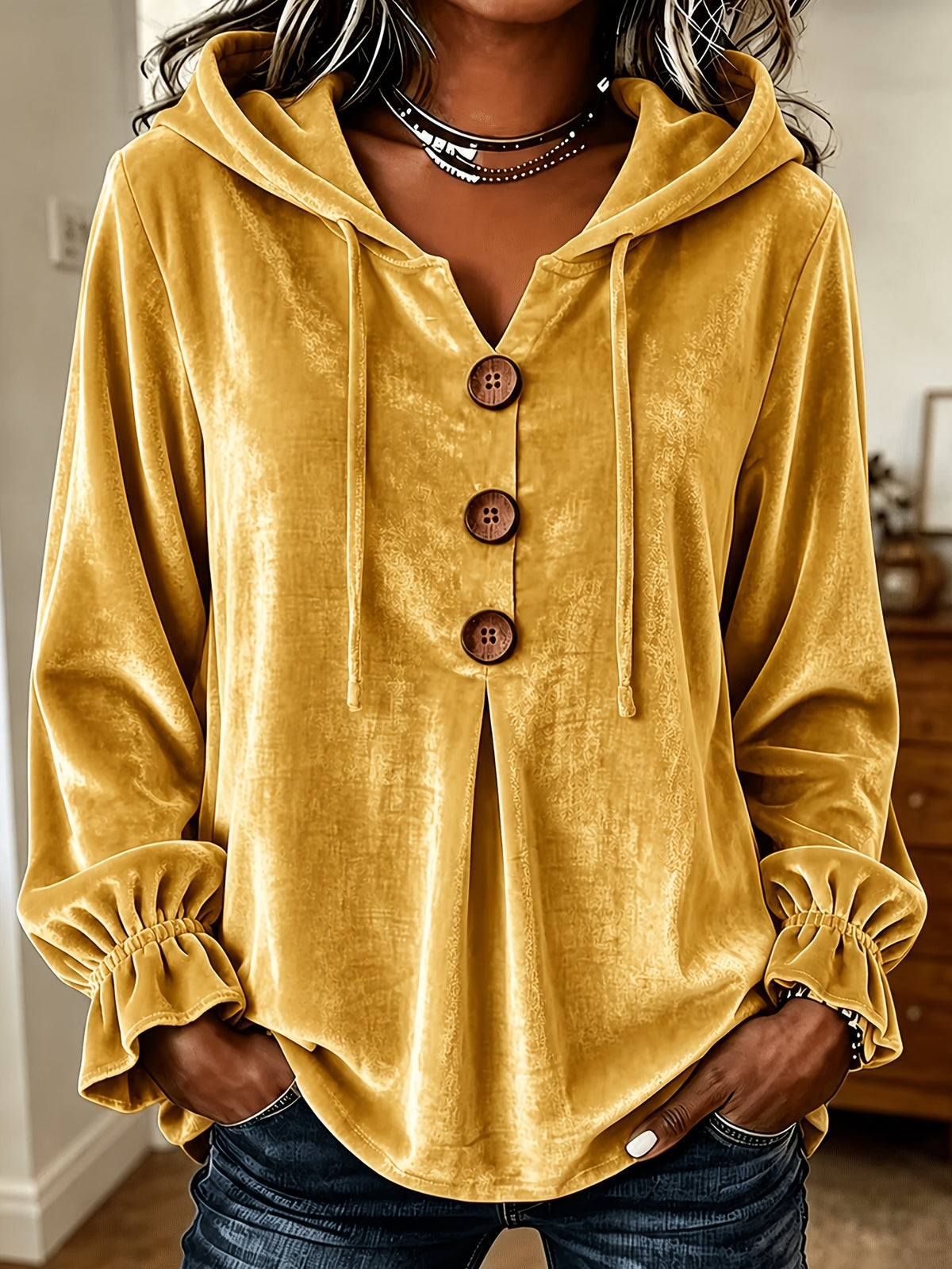 A woman wearing a mustard yellow velvet hooded blouse with a split neckline, three large brown buttons, and gathered ruffled cuffs, paired with dark blue jeans.