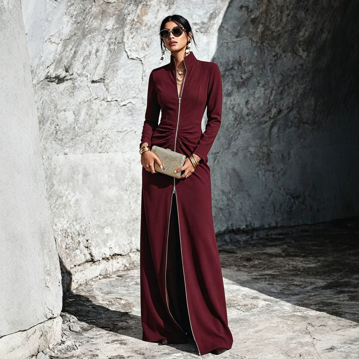 A woman stands outdoors against a textured stone wall wearing a long-sleeve, high-neck, floor-length burgundy dress with a front zipper and side slit. She is holding a metallic clutch and wearing sunglasses and jewelry.
