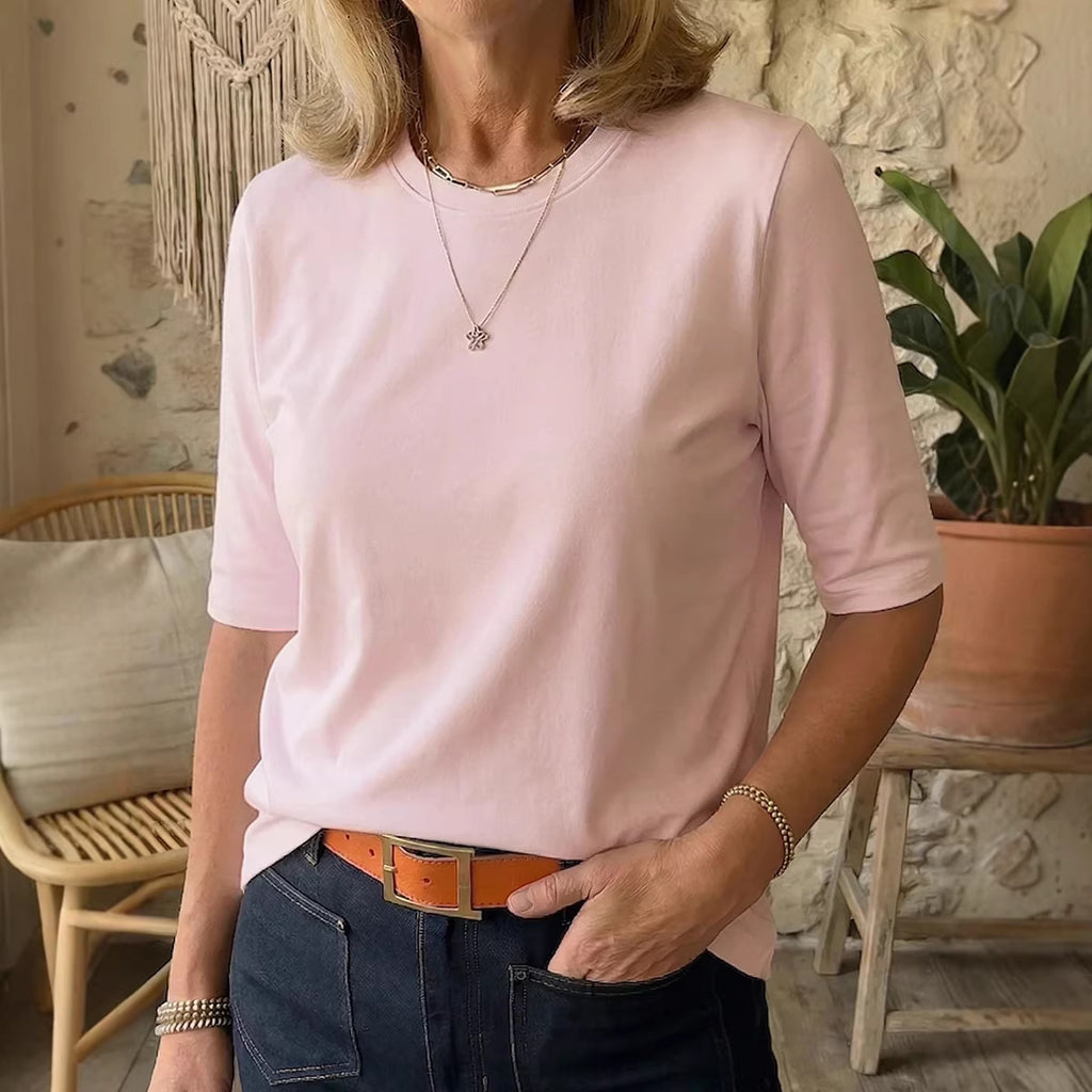 A woman wearing a light pink short-sleeve crewneck t-shirt, styled with dark blue jeans and a brown belt, in a cozy indoor setting with a stone wall and a potted plant.