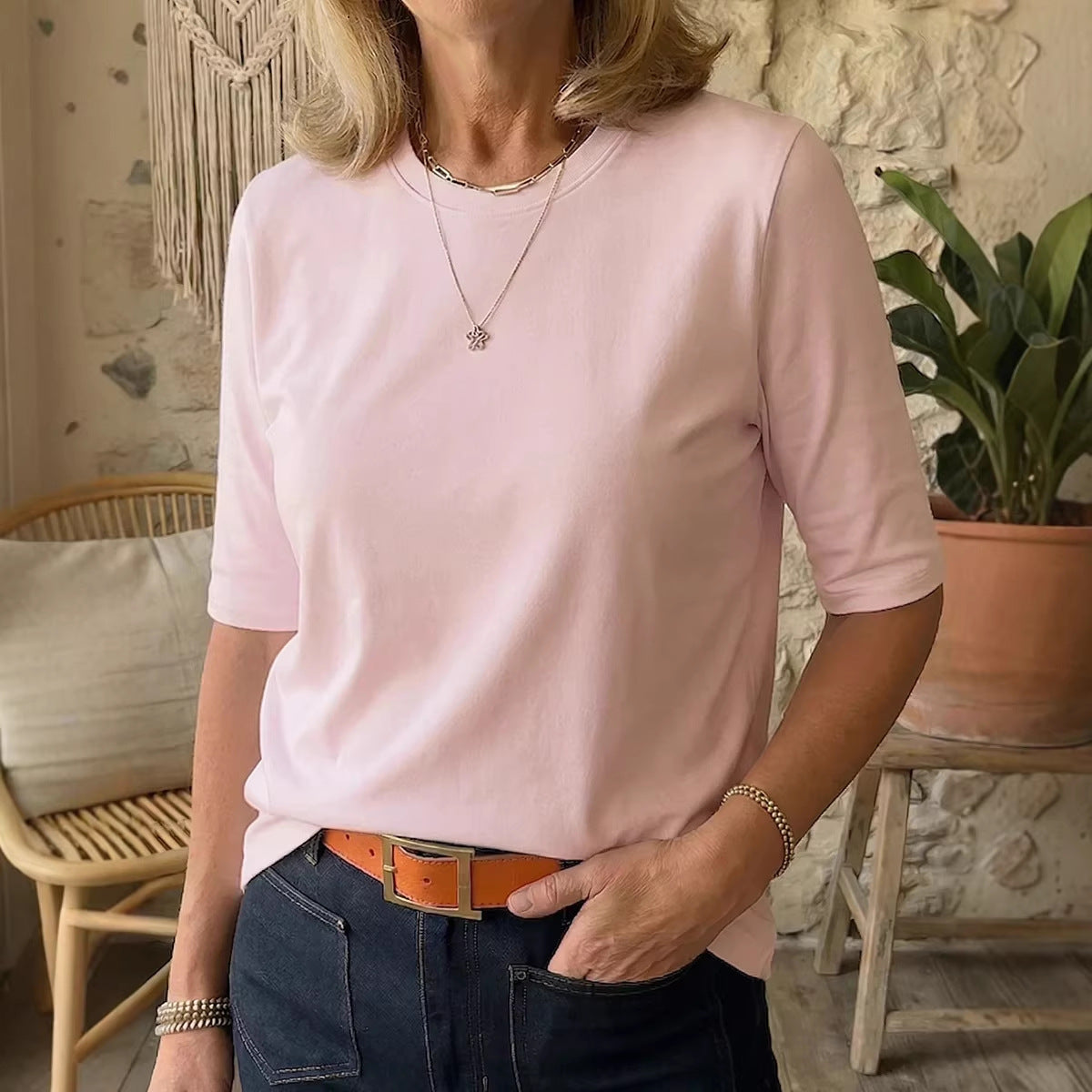 A woman wearing a light pink short-sleeve crewneck t-shirt, styled with dark blue jeans and a brown belt, in a cozy indoor setting with a stone wall and a potted plant.