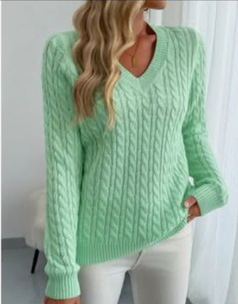 A woman wearing a mint green cable-knit sweater with a V-neck and white pants, standing indoors near a white curtain.