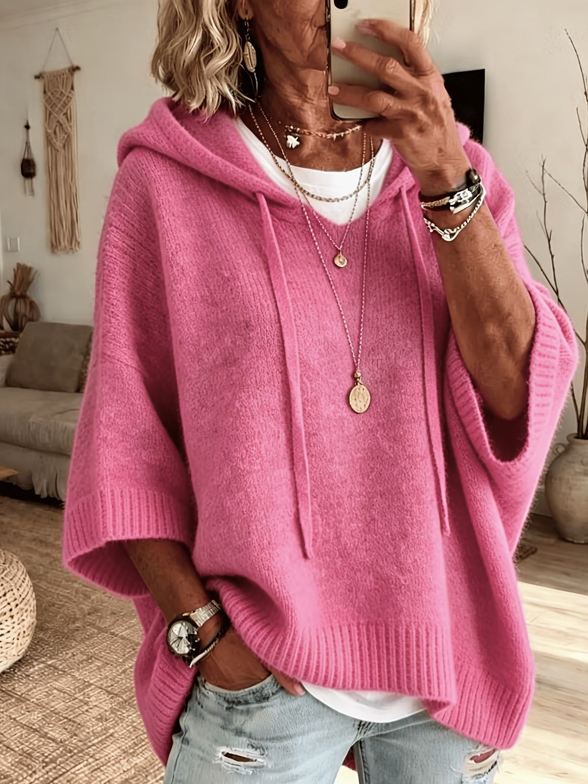 A woman takes a mirror selfie wearing a bright pink knit hooded poncho sweater over a white undershirt and light blue distressed jeans. The background shows a cozy living room setting with a couch and wall decor.