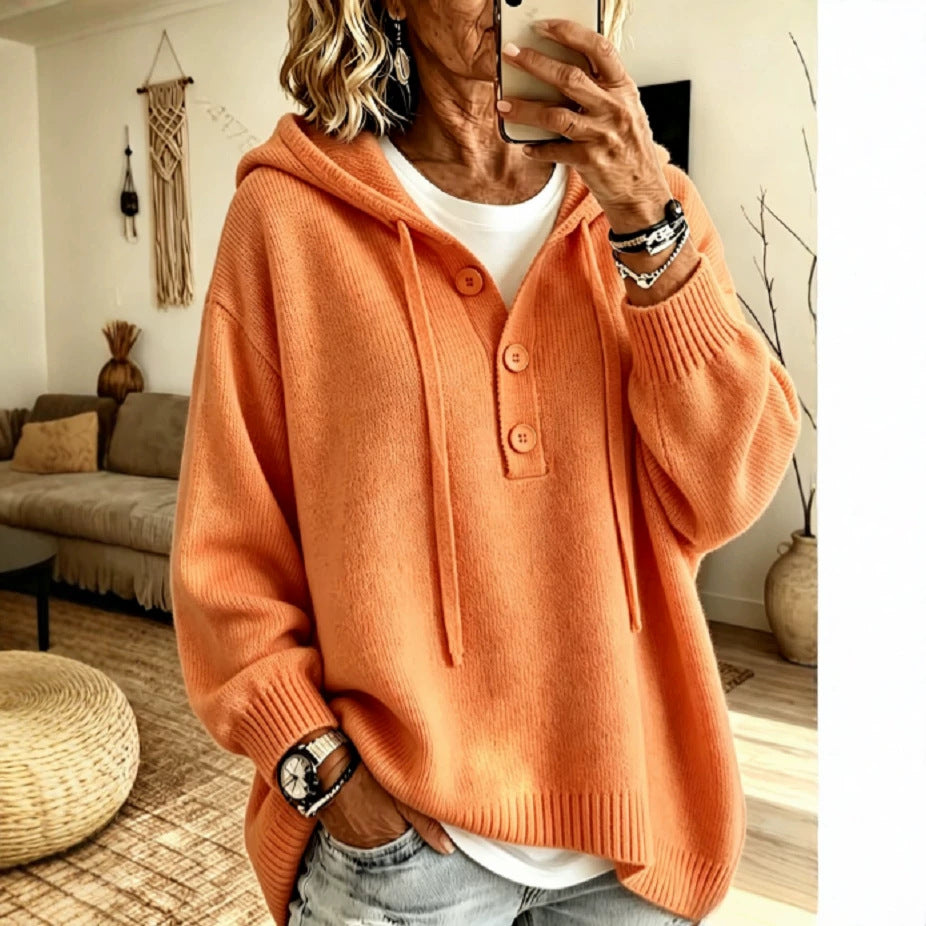 A woman takes a mirror selfie wearing an oversized orange knit hoodie with a buttoned placket over a white top and light-wash jeans, in a cozy living room setting.