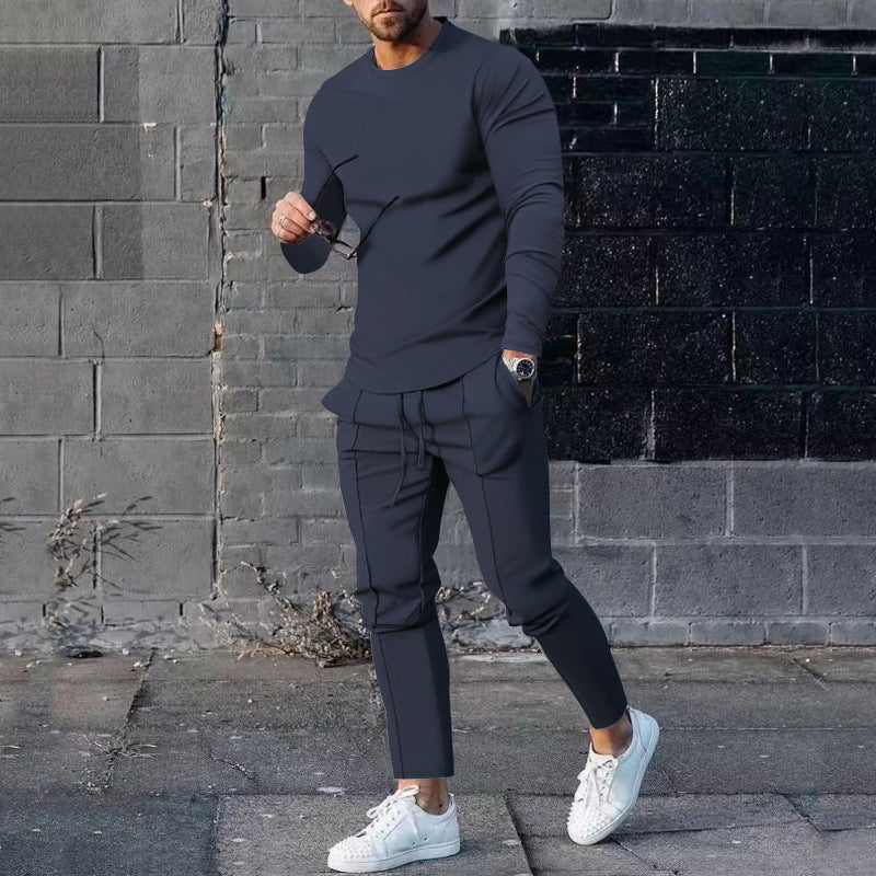 Man wearing a dark gray outfit with white sneakers against a brick wall.