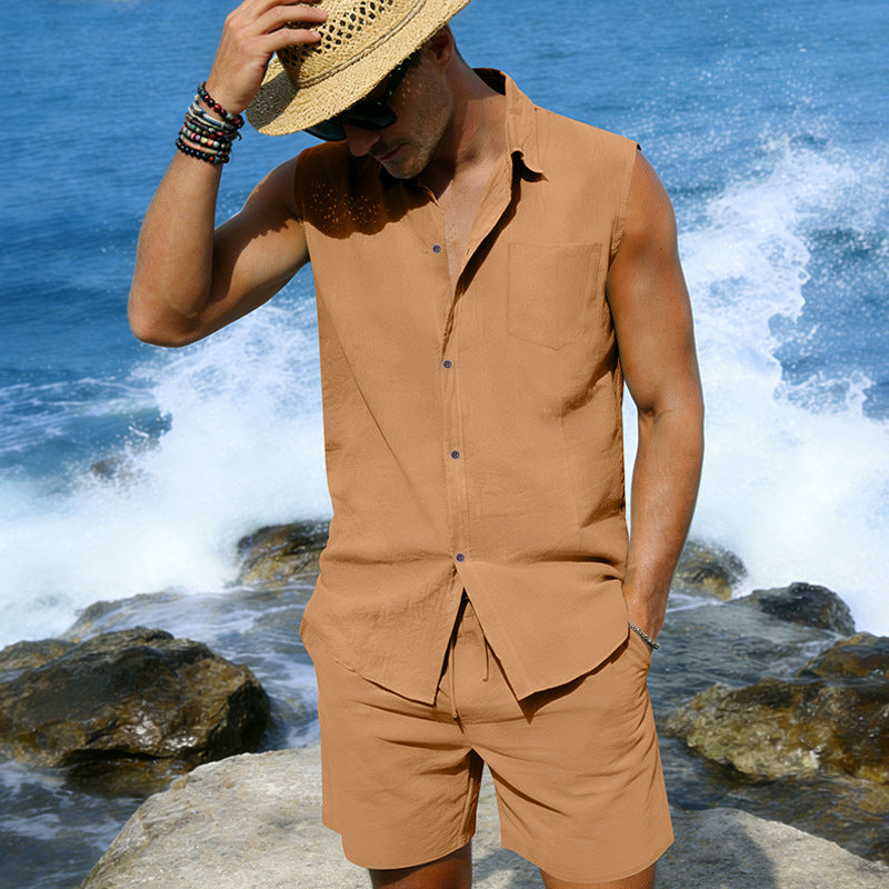 Man in a tan sleeveless shirt and shorts standing by the ocean with waves crashing around.