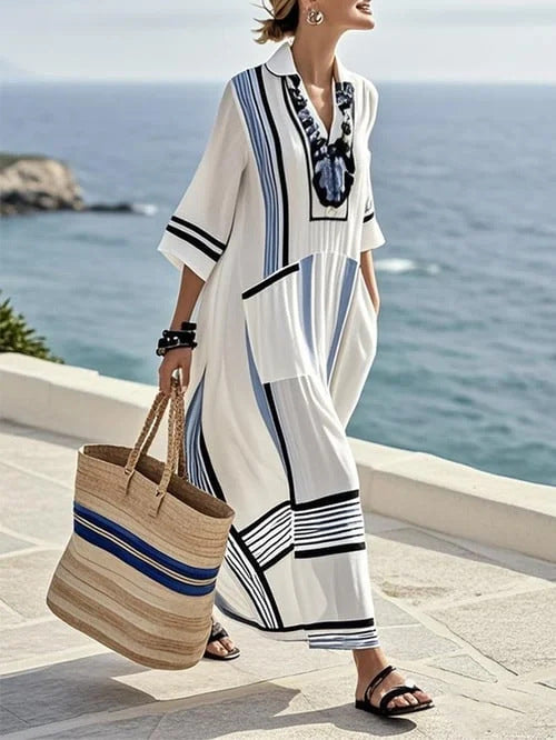 Woman in a long, patterned dress holding a striped bag by the ocean.