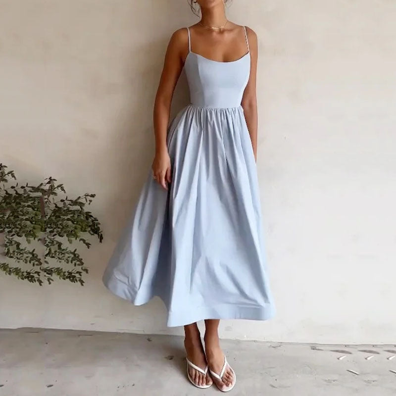 Woman wearing a light blue dress standing against a plain wall with a plant on the left.