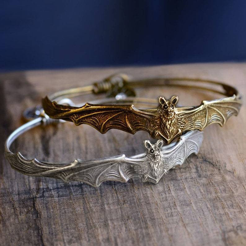 Two bat-shaped bracelets, one gold and one silver, on a wooden surface with a dark background.