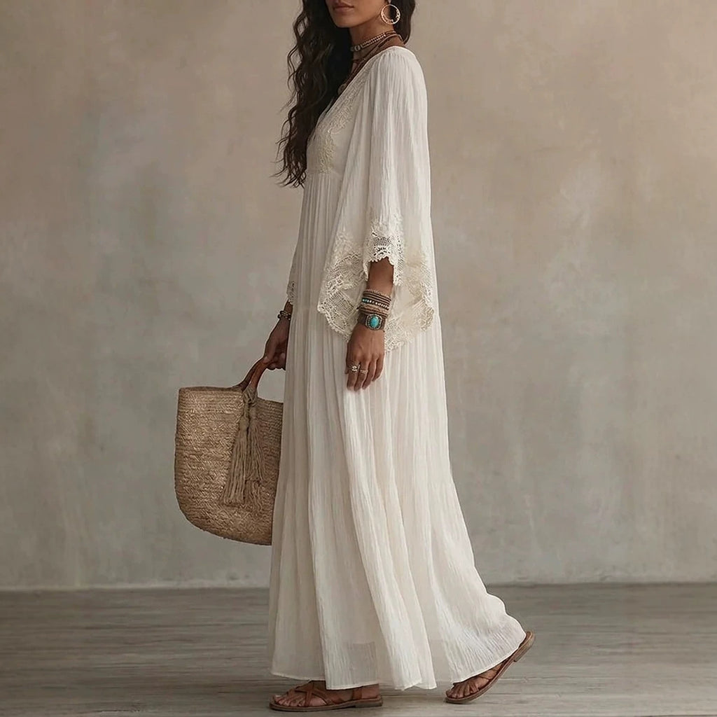 A woman wearing a long white bohemian maxi dress with lace trim on the sleeves, holding a woven straw tote bag, standing against a textured beige wall.