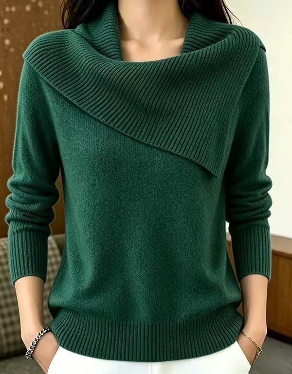 A woman wearing a dark green knit sweater with an asymmetrical draped cowl neck and long sleeves, paired with white pants.