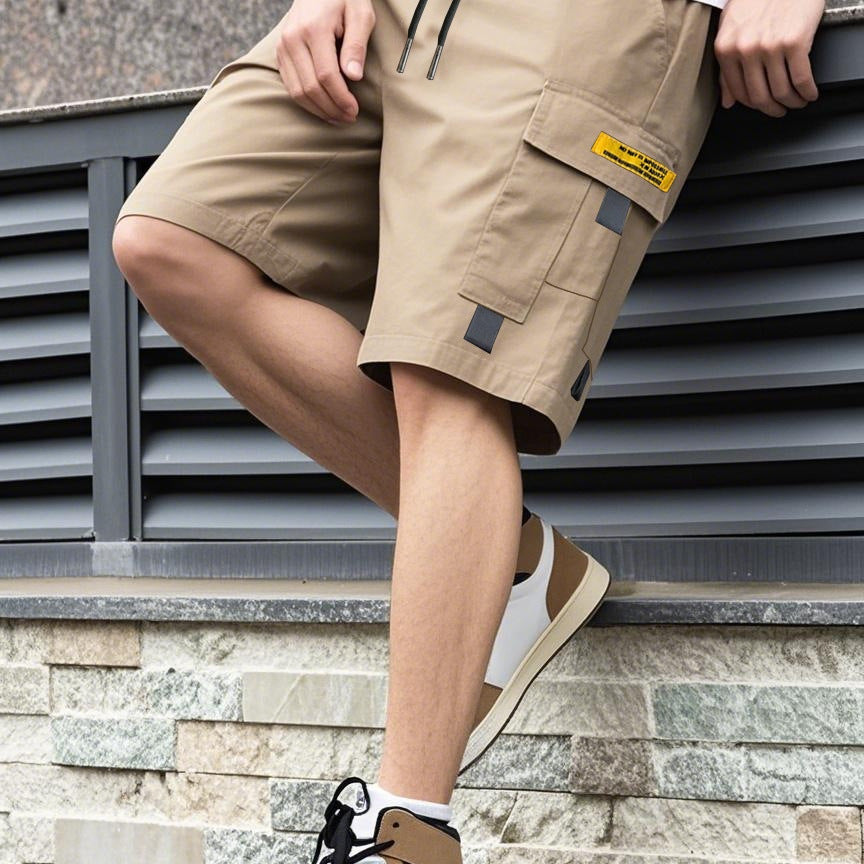 Person wearing beige cargo shorts with a brand label, standing against a stone wall.