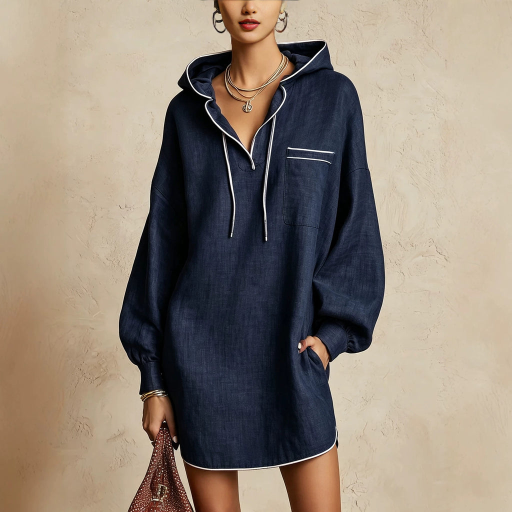 A woman models a navy blue hooded shirt dress with white piping details, standing against a textured beige wall and holding a brown patterned handbag.
