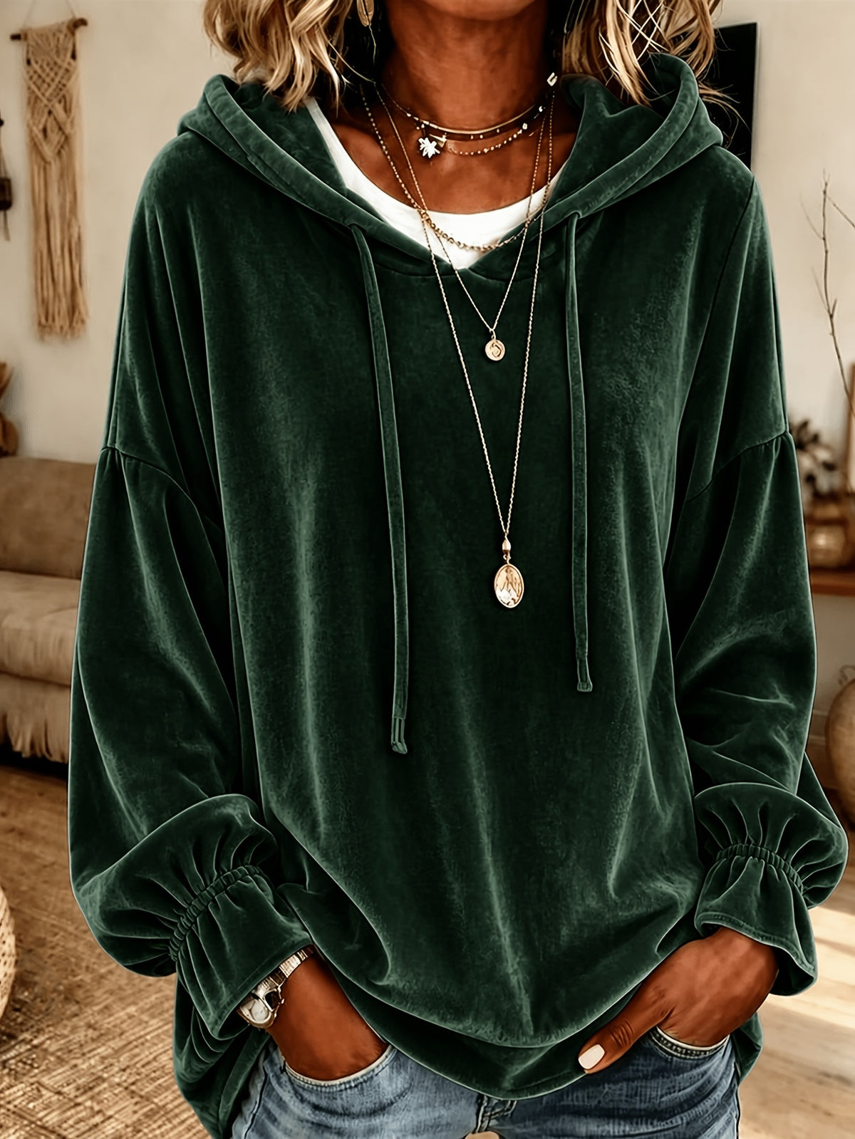 A woman wearing a dark forest green velvet hoodie with drawstrings and elasticized cuffs, layered over a white t-shirt and accessorized with layered necklaces, paired with blue jeans in a cozy indoor setting.
