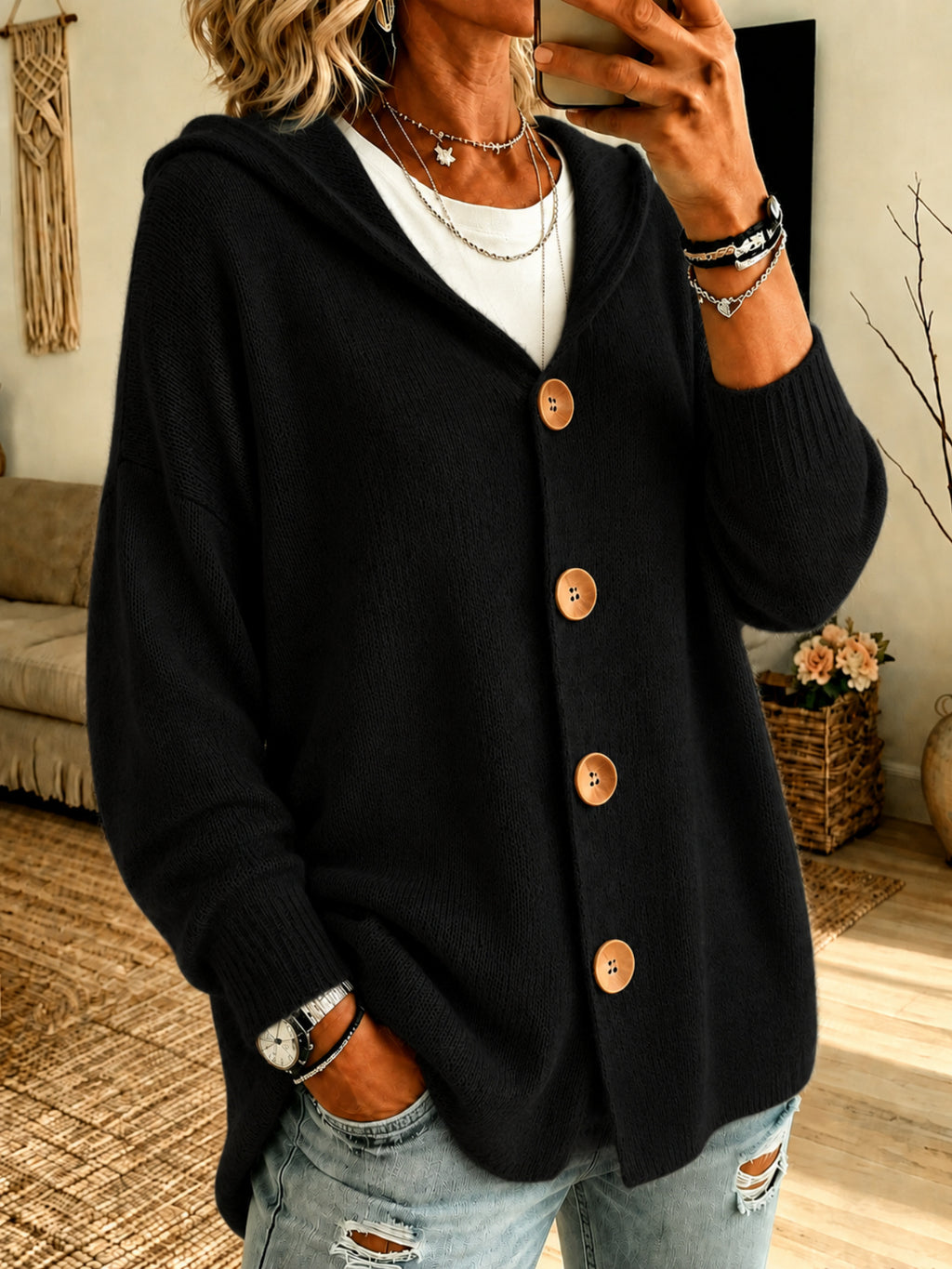 A woman wearing a black knit cardigan with a hood and large brown buttons, layered over a white t-shirt and paired with distressed light blue jeans in a cozy indoor setting.