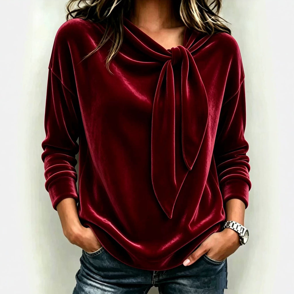 A woman wearing a long-sleeve velvet top in deep burgundy red with a knotted cowl neckline, paired with blue jeans, against a plain light background.