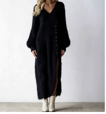 A woman wearing a long black fuzzy knit cardigan dress with a V-neck, balloon sleeves, and a side button closure, paired with off-white ankle boots, set against a plain light gray wall.