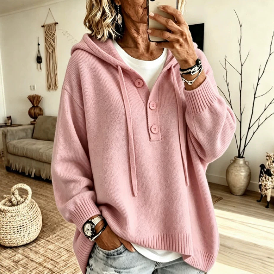 A woman takes a mirror selfie wearing a pink knit hooded sweater with a buttoned placket over a white top and light-wash jeans, in a cozy living room setting.