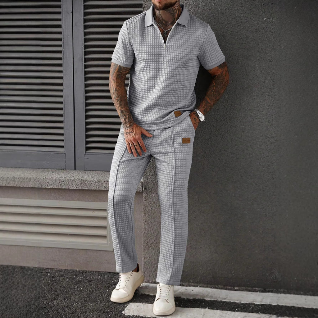 Man wearing a checkered shirt and pants with white sneakers against a gray wall.