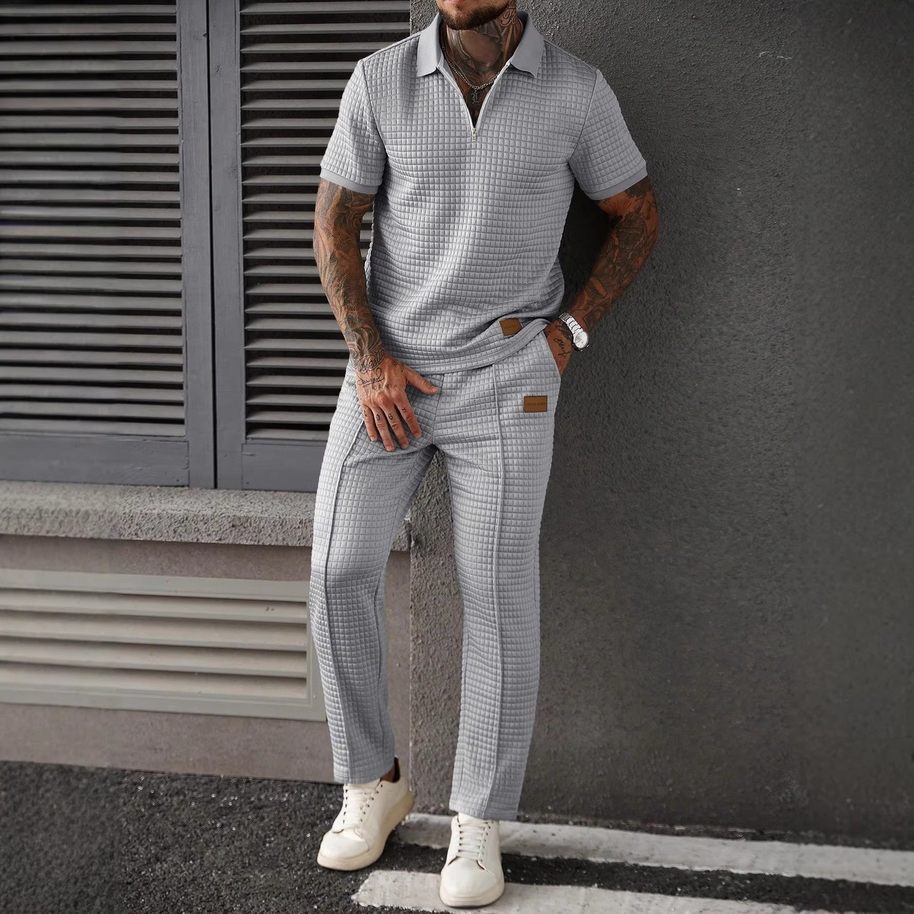 Man wearing a checkered shirt and pants with white sneakers against a gray wall.
