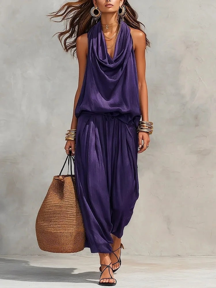 Woman wearing a black sleeveless top and harem pants, holding a woven bag against a neutral background.