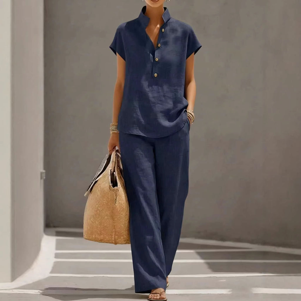 Person wearing a navy outfit with wide-leg pants and holding a beige bag against a gray background