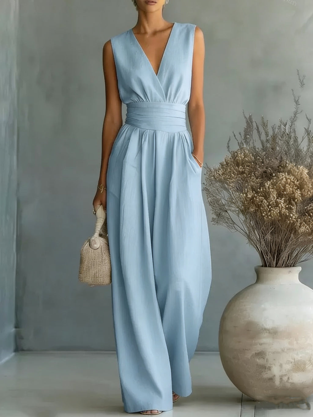 Woman wearing a light blue dress standing next to a vase with dried flowers against a gray wall.