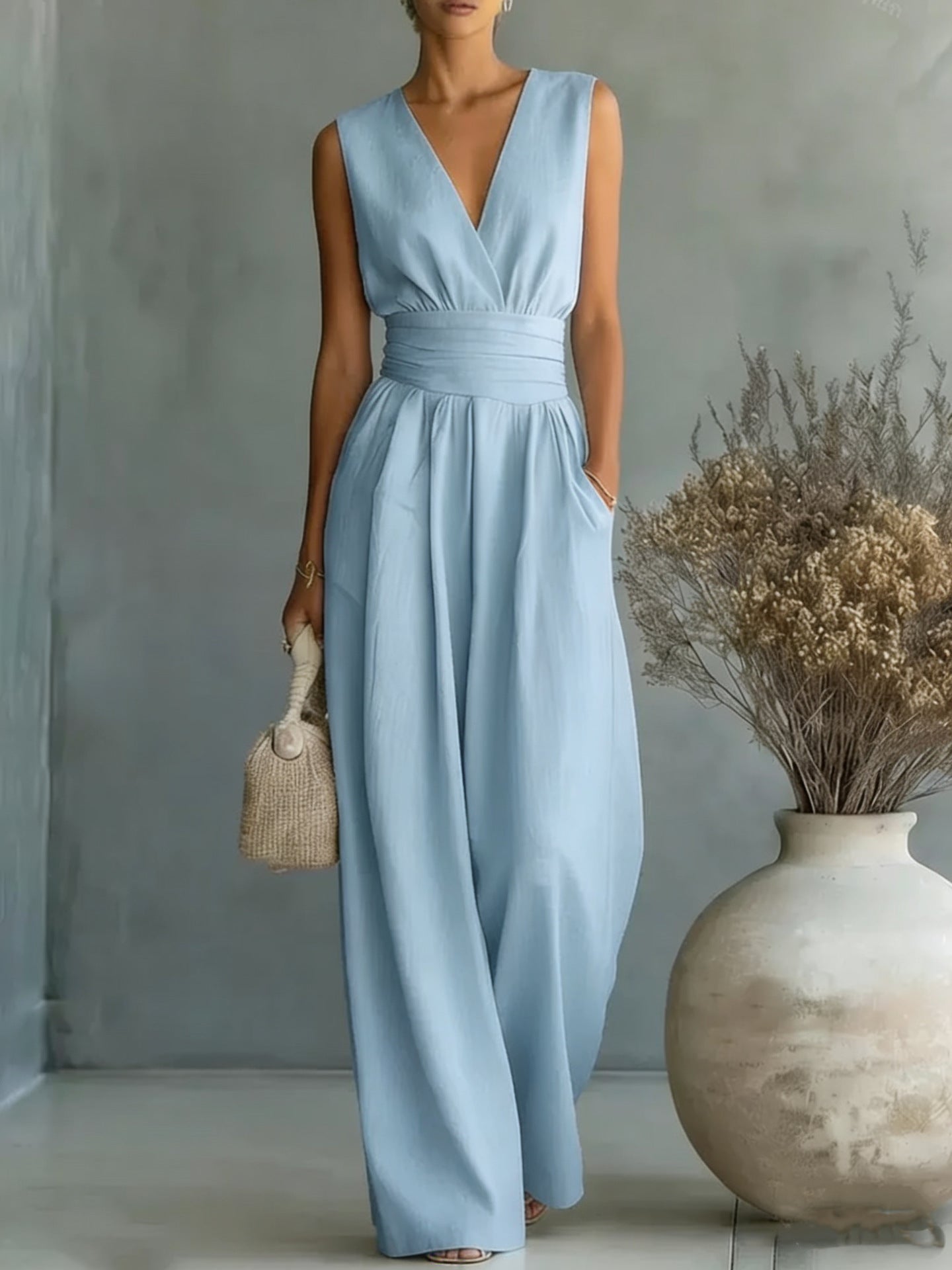 Woman wearing a light blue dress standing next to a vase with dried flowers against a gray wall.