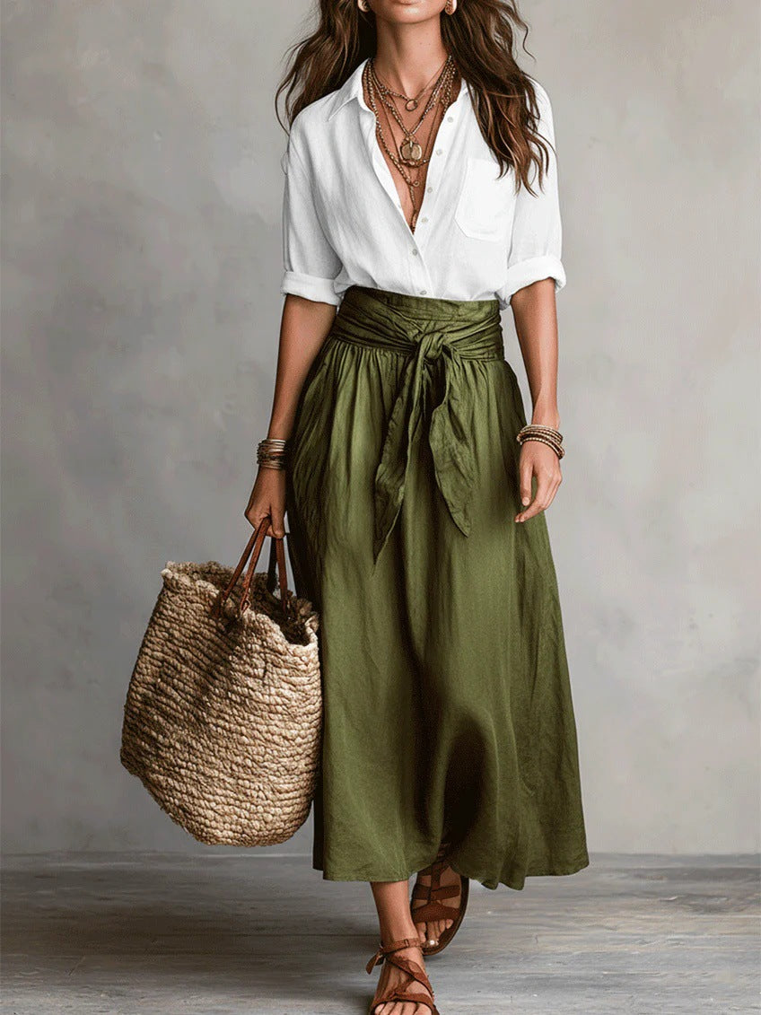 Woman wearing a white blouse and green skirt holding a woven bag against a gray background