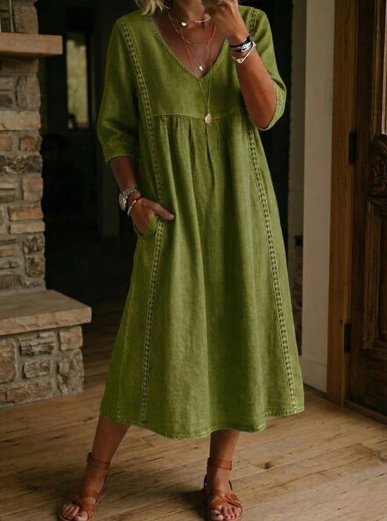 A woman wearing a loose olive green midi dress with three-quarter sleeves, a V-neckline, and decorative vertical stitching details, standing on a wooden floor in an indoor setting.