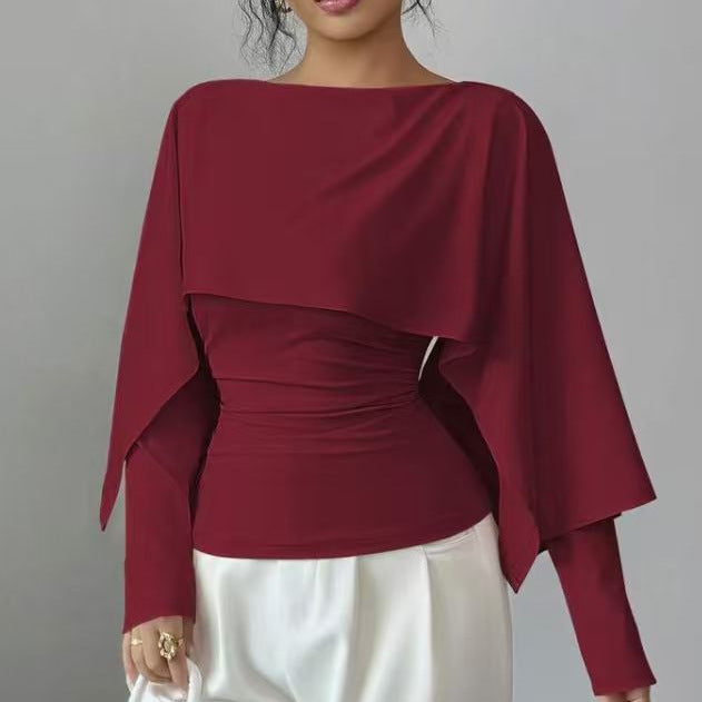 A woman wearing a deep burgundy long-sleeve top with a draped, asymmetrical layered overlay and gathered waist detail, paired with white pants, against a plain gray background.