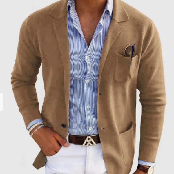 Man wearing a brown blazer over a blue striped shirt with white pants on a light gray background