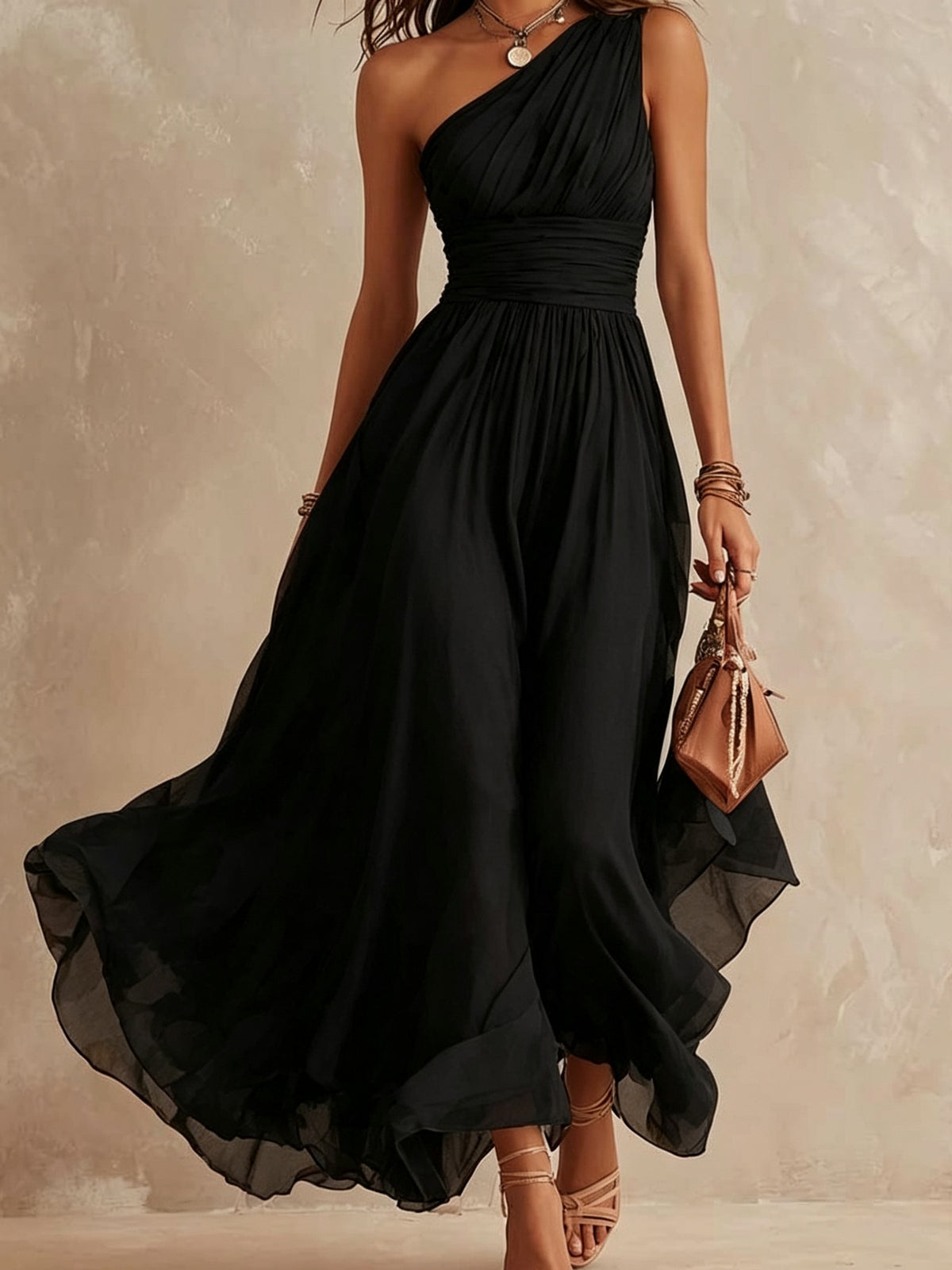 A woman wearing a flowing black one-shoulder maxi dress with a gathered waist, holding a small brown handbag against a textured beige studio backdrop.