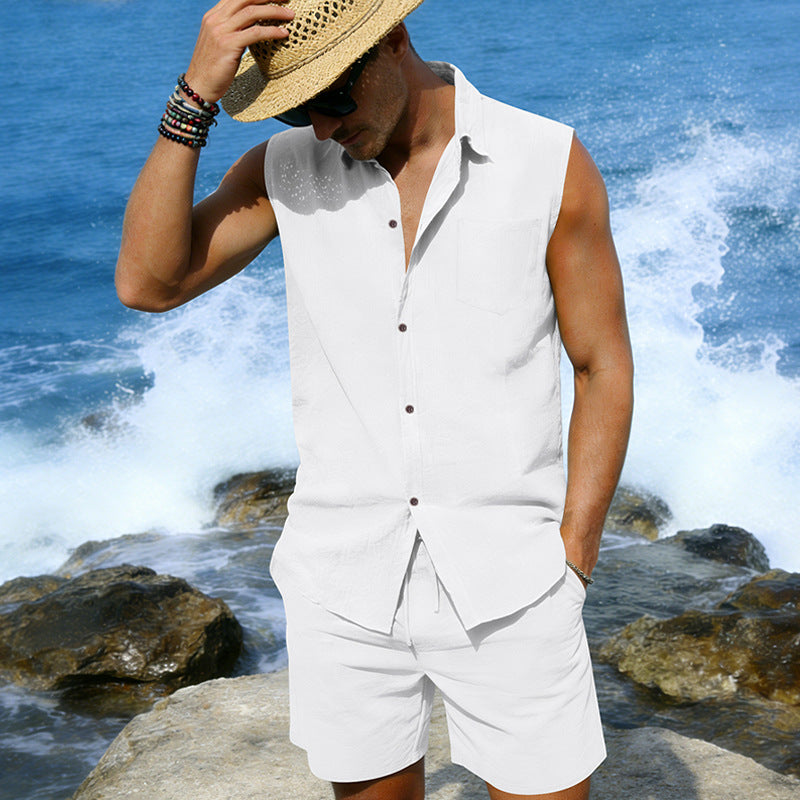 Man in a white sleeveless shirt and shorts standing by the ocean with waves crashing around.
