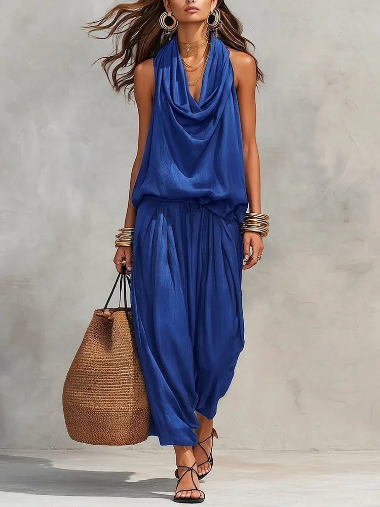 Woman wearing a blue sleeveless top and harem pants, holding a woven bag against a neutral background.