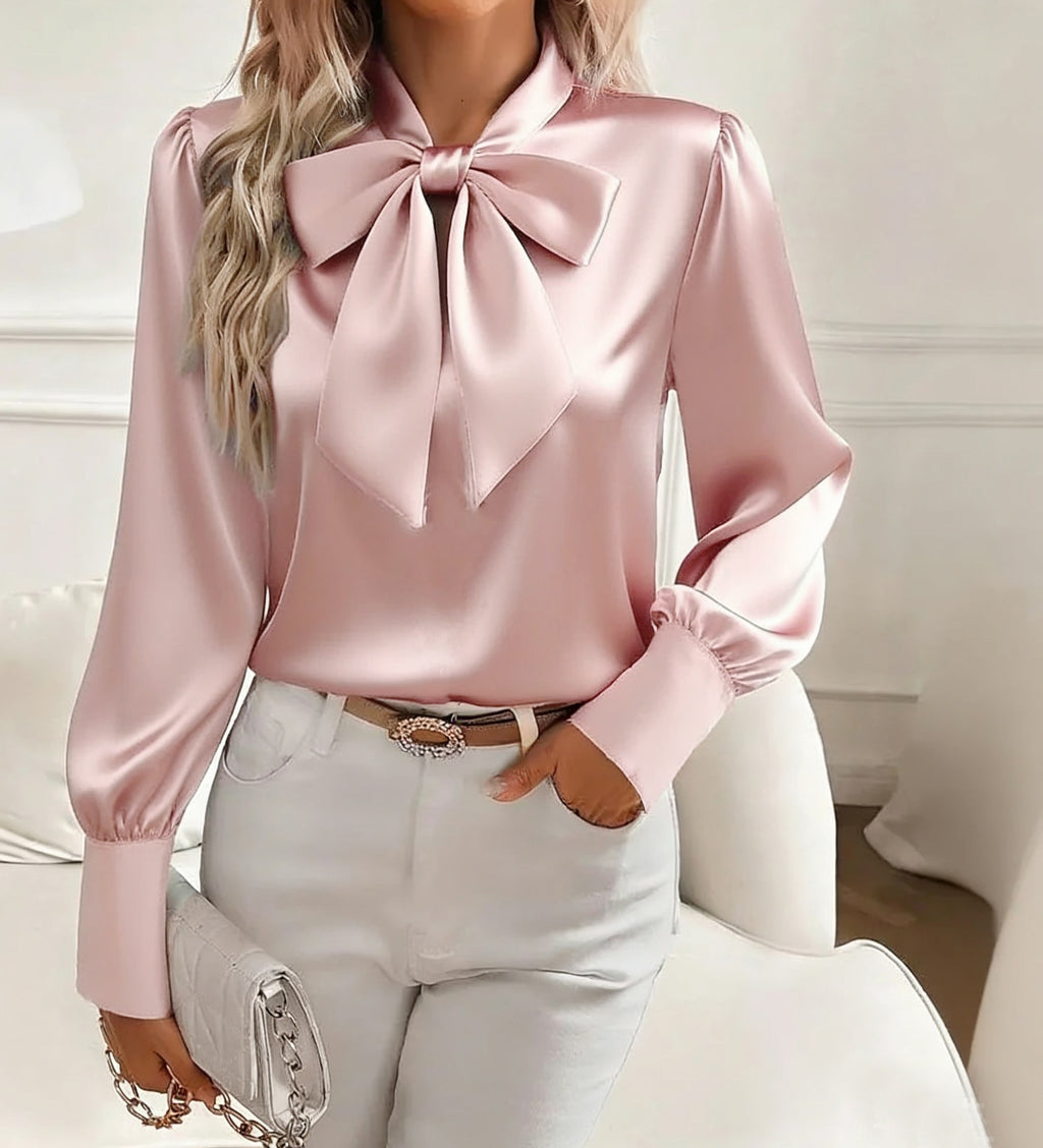 Pink satin blouse with a large bow and white pants on a neutral background