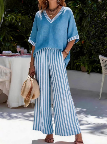 Woman wearing a blue top and striped pants outdoors with a table and chairs in the background.
