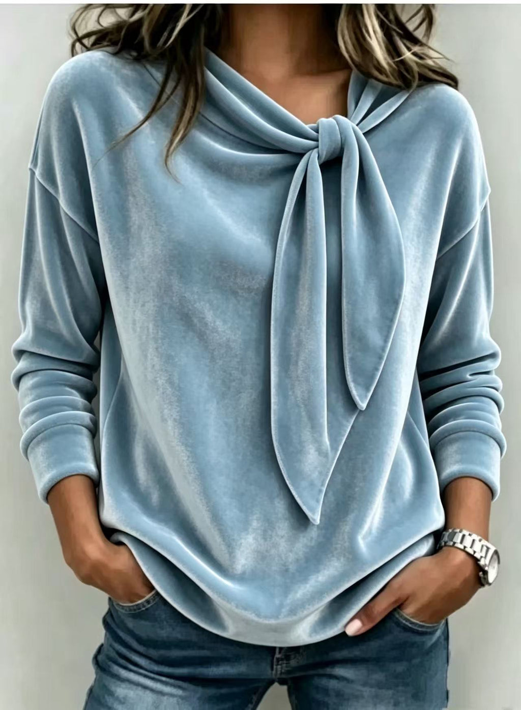 A woman wearing a light blue long-sleeve velvet top with a large front knot detail, paired with blue jeans.