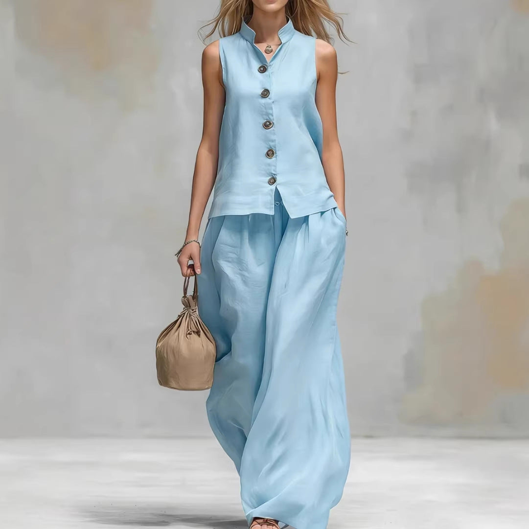 Woman wearing a light blue sleeveless top and wide-leg pants, holding a beige handbag against a neutral background.