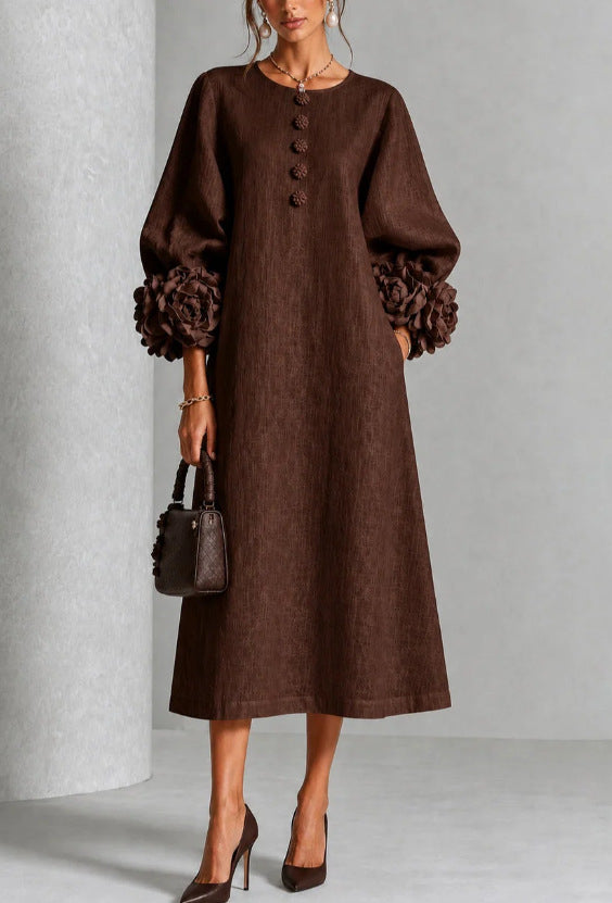 A woman models a brown midi dress with voluminous long sleeves featuring textured pom-pom cuffs, a buttoned round neckline, and side pockets. She is holding a matching brown handbag and wearing brown high heels against a neutral gray studio background.