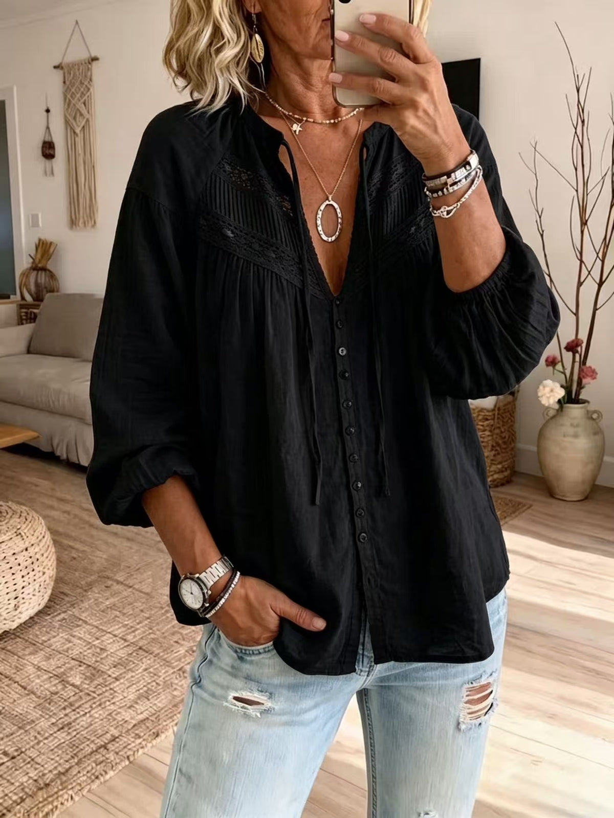 A woman wearing a black long-sleeve button-up blouse with a deep V-neck and embroidered detailing on the chest, paired with light-wash distressed jeans, shown in a mirror selfie inside a living room.
