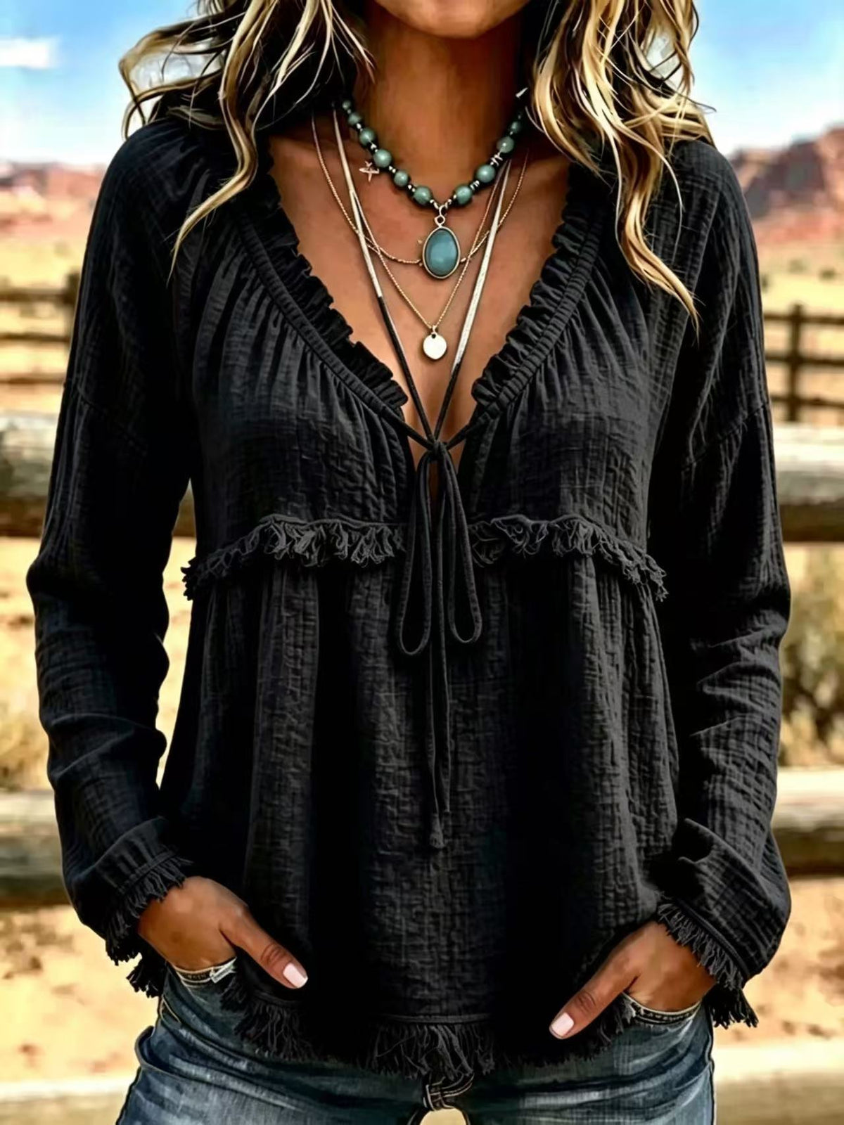 A woman wearing a black long-sleeve crinkle gauze top with a ruffled V-neckline, tie-front detail, and frayed hem, paired with layered necklaces and jeans, set against a blurred outdoor desert background.