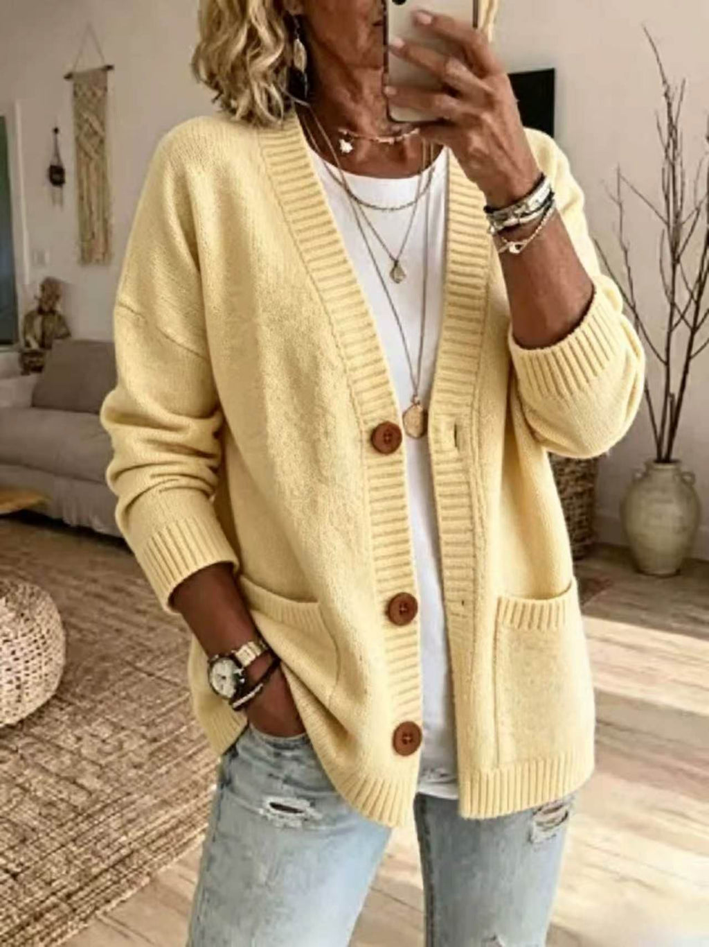 A woman wearing a light yellow knit cardigan with brown buttons over a white top and light blue jeans, taking a mirror selfie in a cozy living room setting.