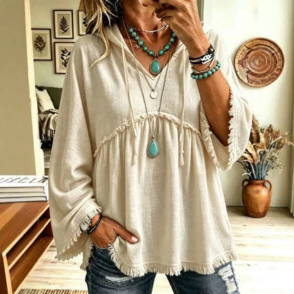 A woman wearing a cream-colored bohemian-style tunic top with fringe details, paired with distressed blue jeans and layered turquoise and silver necklaces, in a cozy indoor setting.