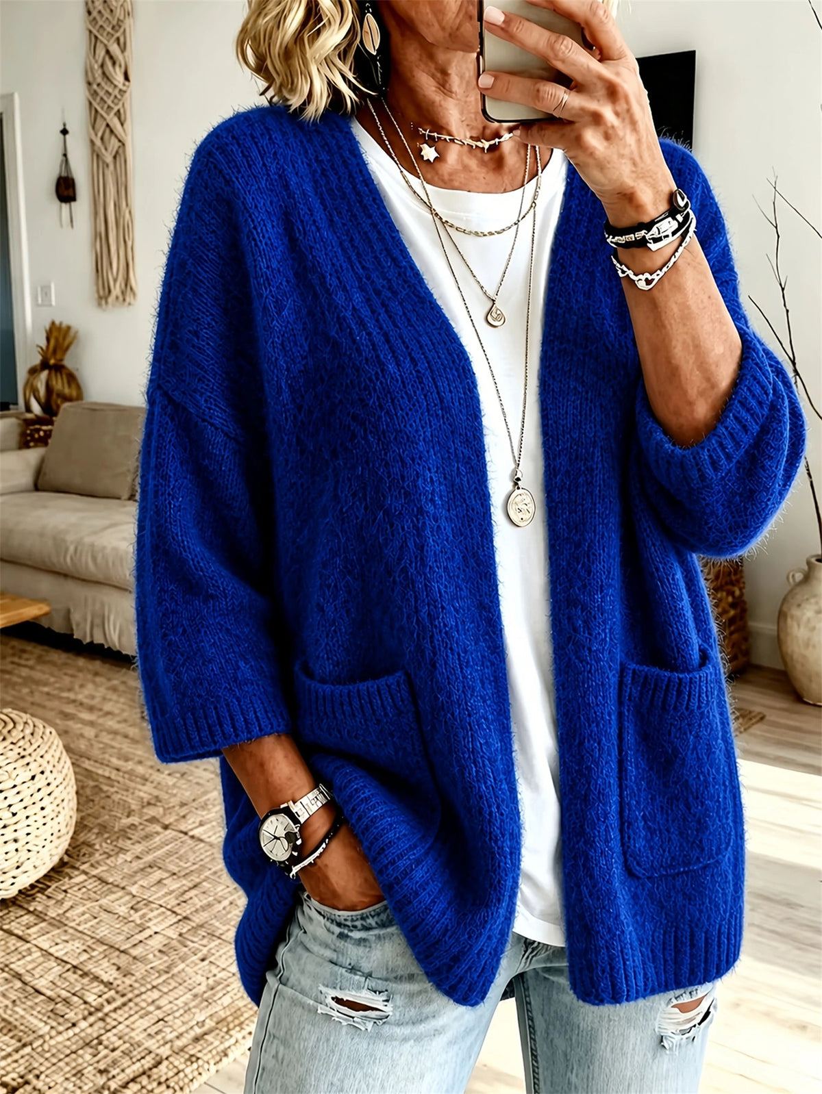 A woman wearing a vibrant royal blue chunky knit cardigan with front pockets over a white t-shirt and distressed light-wash jeans, taking a mirror selfie in a cozy living room setting.