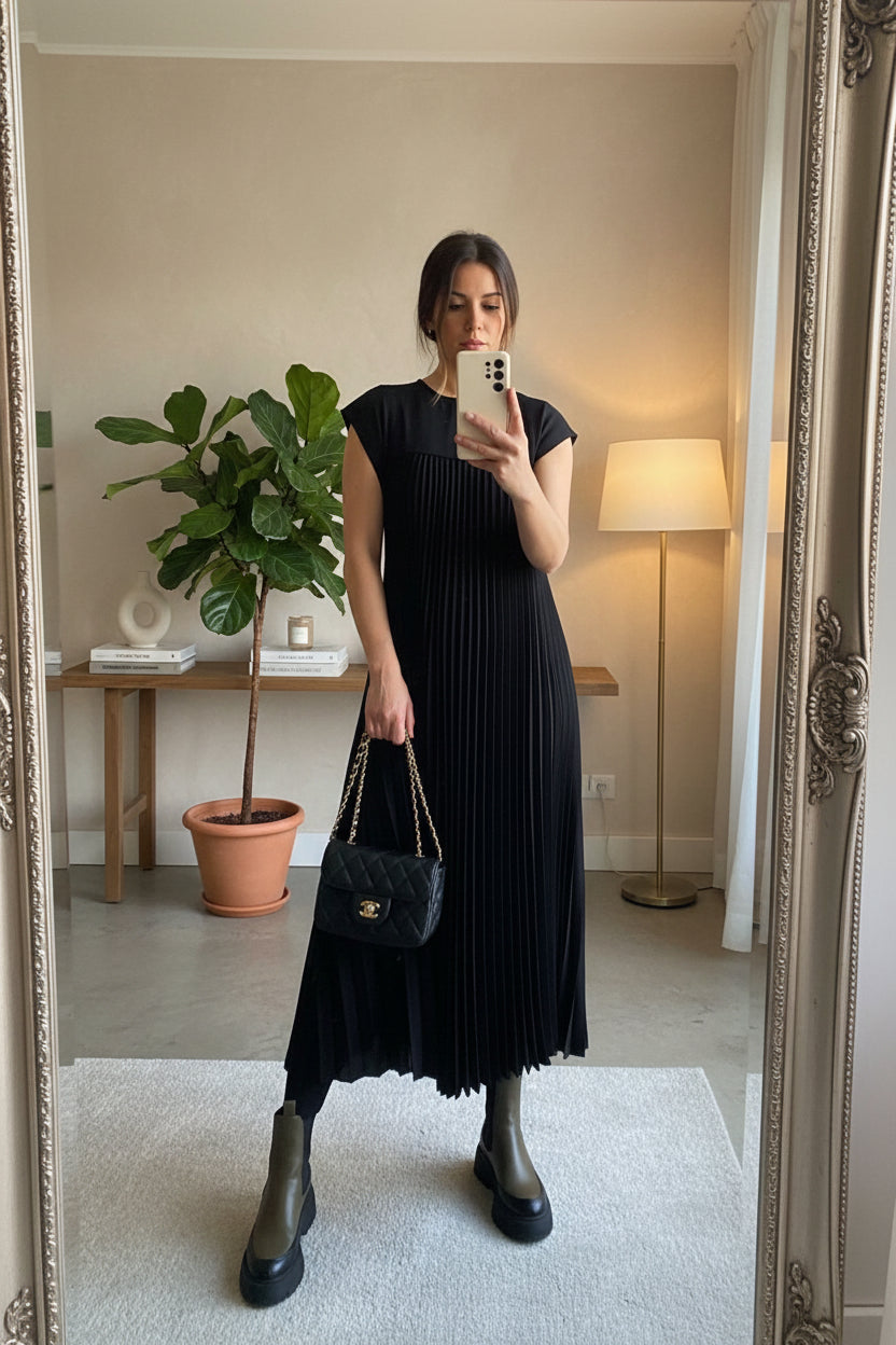 Person wearing a black pleated dress holding a black handbag in a room with decor elements.